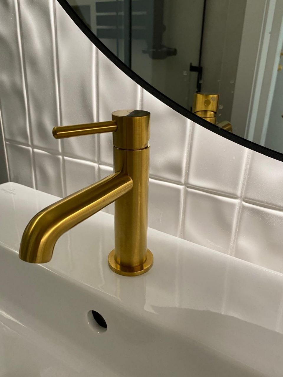 Błyszcząca, złota bateria umywalkowa w nowoczesnej łazience z białą umywalką i płytkami. Widoczne odbicie baterii w okrągłym lustrze.