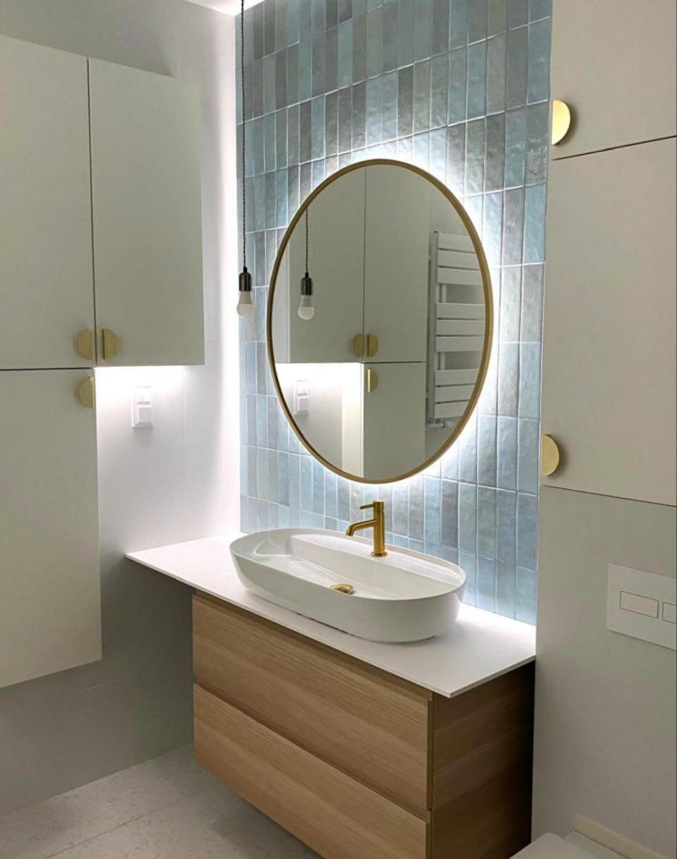 Stylowa łazienka z owalnym lustrem podświetlanym LED, umywalką nablatową i błękitnymi płytkami ułożonymi pionowo. Złote akcenty armatury i oświetlenia.