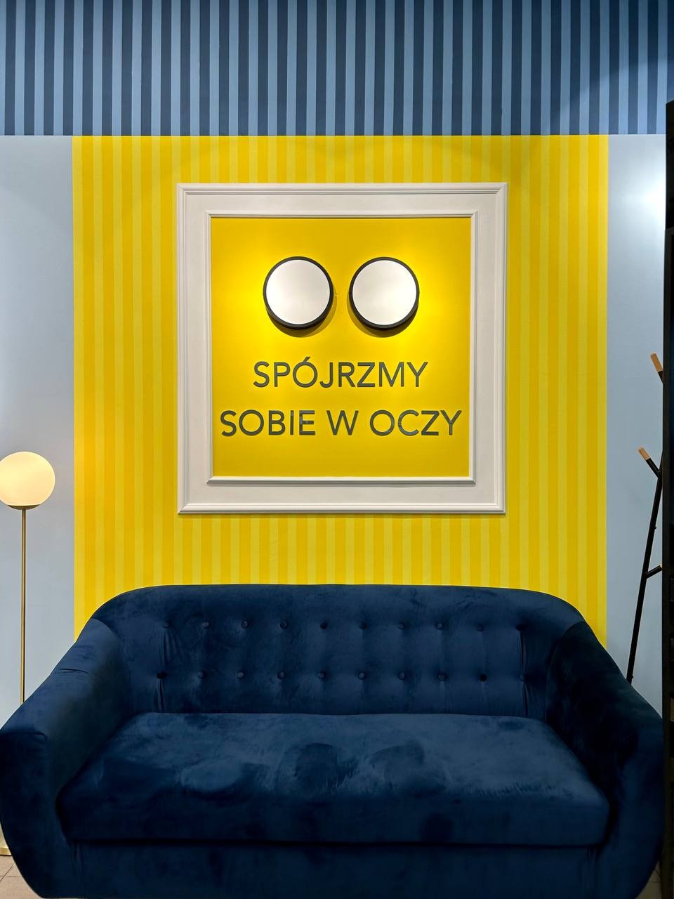 Niebieska sofa na tle żółtej ściany w paski z oprawionym napisem 'Spójrzmy sobie w oczy' i okrągłymi lampami. Nowoczesne wykończenie wnętrza.