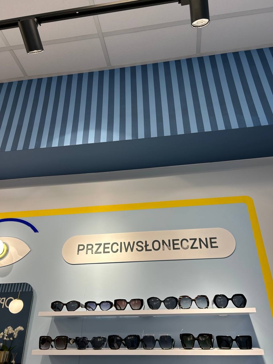 Półki z okularami przeciwsłonecznymi w salonie optycznym, podświetlone lampami sufitowymi. Ściana w kolorze błękitu z żółtymi akcentami i napisem 'Przeciwsłoneczne'.