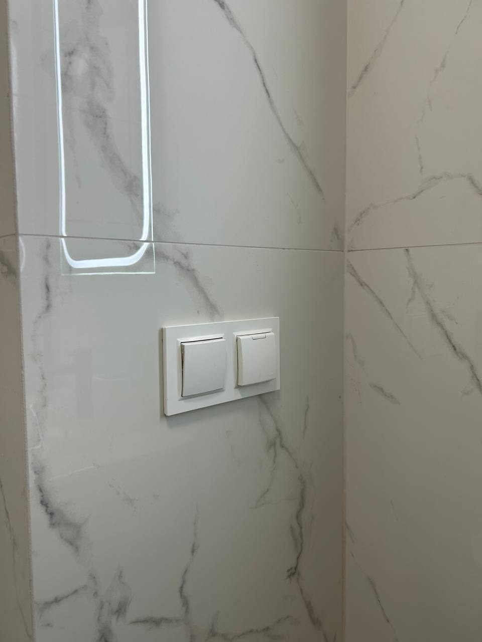 Białe, kwadratowe włączniki światła na ścianie wyłożonej błyszczącymi, marmurkowymi płytkami w łazience. Minimalistyczny design.