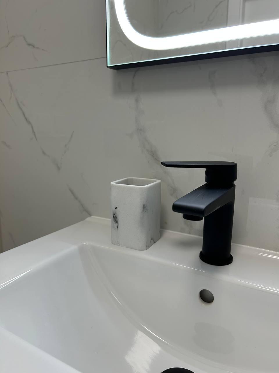 Biała umywalka z czarną armaturą i marmurową ścianą w tle, oświetlone lustro. Detal łazienki po remoncie w Gdańsku. Nowoczesny design.
