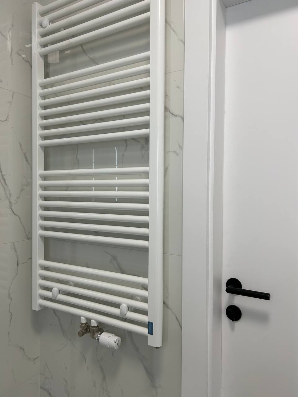 Biały grzejnik drabinkowy na marmurowej ścianie w łazience, obok białe drzwi z czarną klamką. Minimalistyczny design, nowoczesne wykończenie.