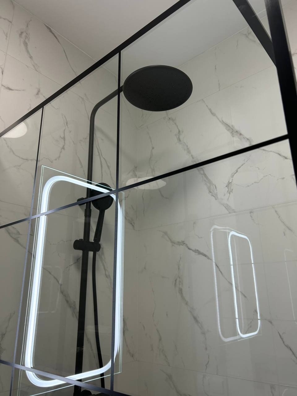 Nowoczesna kabina prysznicowa z czarnymi profilami i deszczownicą na tle marmurowych płytek. Widoczne odbicie oświetlenia w szkle.