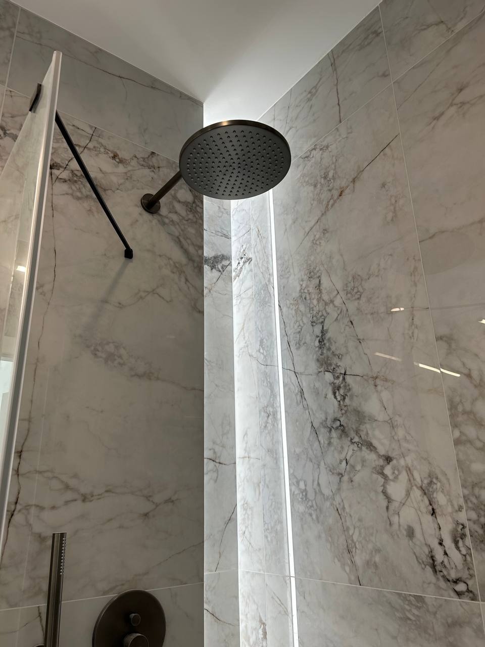 Nowoczesna kabina prysznicowa z marmurowymi płytkami, deszczownicą w kolorze ciemnego brązu i subtelnym oświetleniem LED w pionowej szczelinie między płytkami.