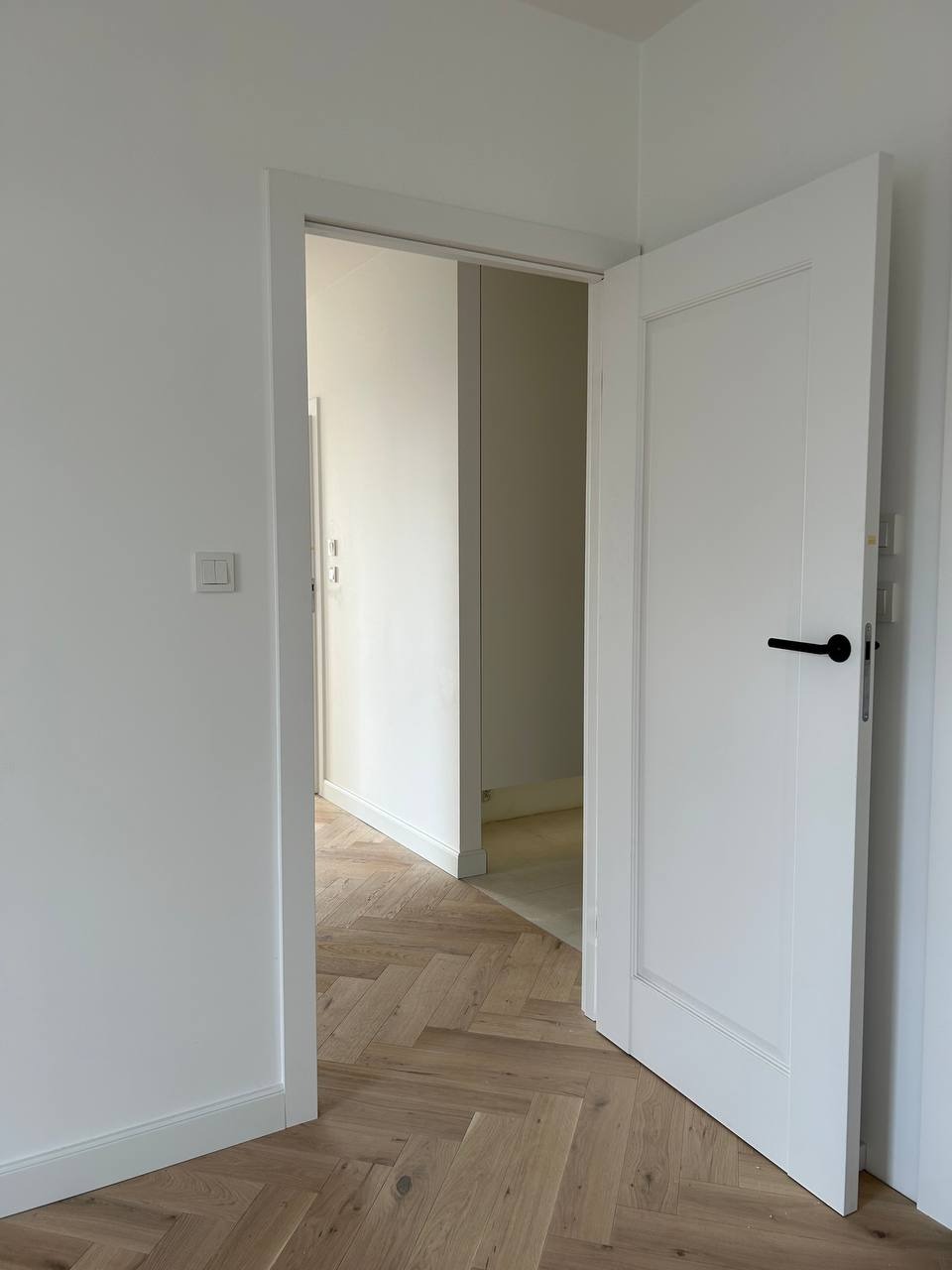 Białe drzwi wewnętrzne z czarną klamką, otwarte na parkiet ułożony w jodełkę. Widok na sąsiednie pomieszczenie z białymi ścianami i listwami przypodłogowymi.