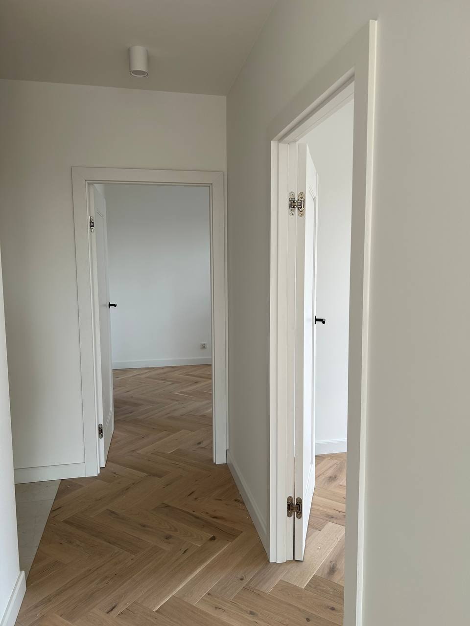 Jasne wnętrze z parkietem w jodełkę, widok na otwarte, białe drzwi. Minimalistyczny design, neutralna kolorystyka, subtelne oświetlenie punktowe.