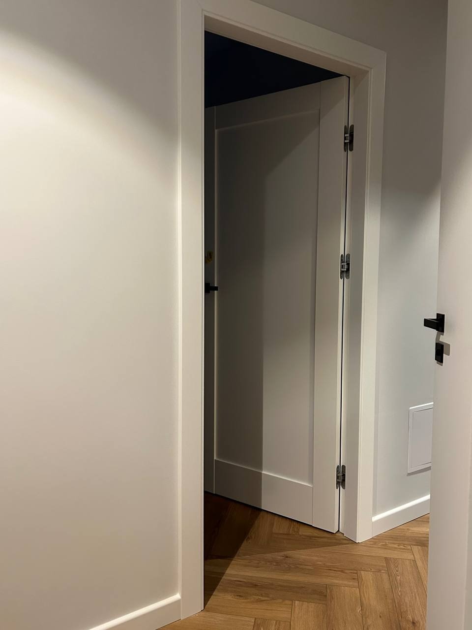 Otwarta, biała, wewnętrzna stolarka drzwiowa z widocznymi zawiasami, na tle jasnych ścian i parkietu ułożonego w jodełkę. Minimalistyczny design.