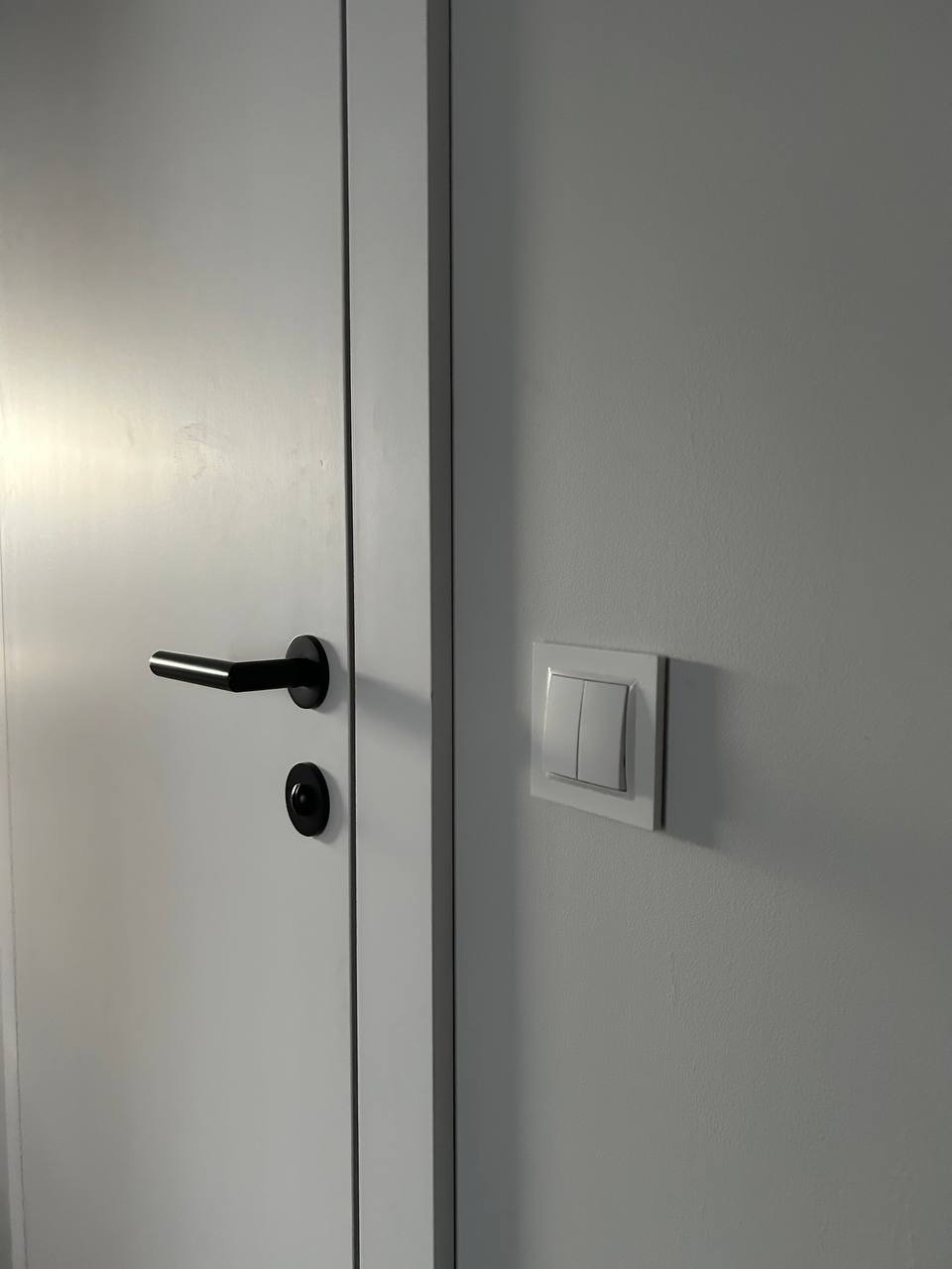 Białe drzwi z czarną klamką i włącznik światła na ścianie. Minimalistyczny design, neutralna kolorystyka, nowoczesne wykończenie. Detal wnętrza po remoncie.