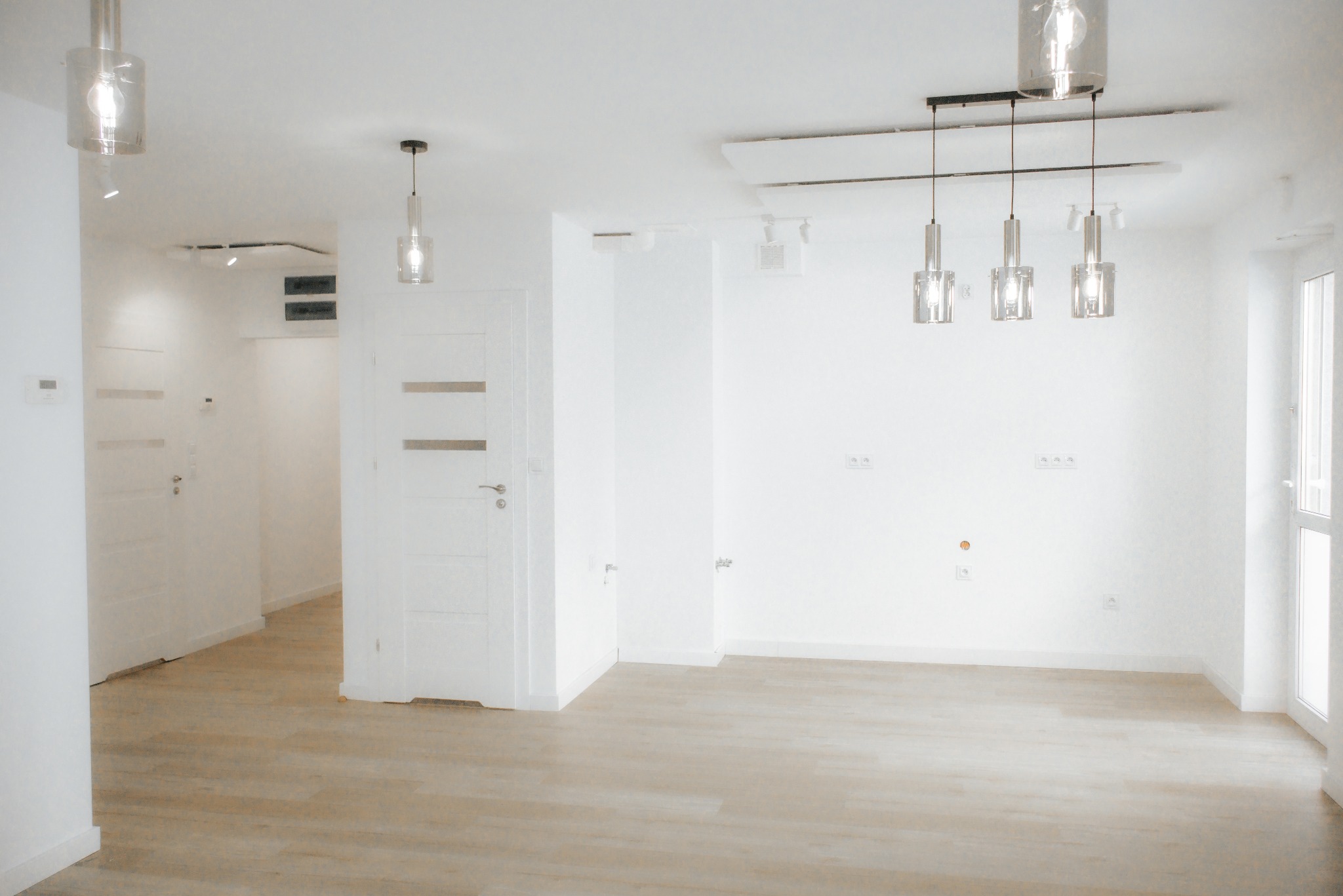Jasne, przestronne wnętrze po remoncie: parkiet, białe ściany, nowoczesne lampy wiszące. Widoczne gniazdka elektryczne i drzwi wewnętrzne. Minimalistyczny design.