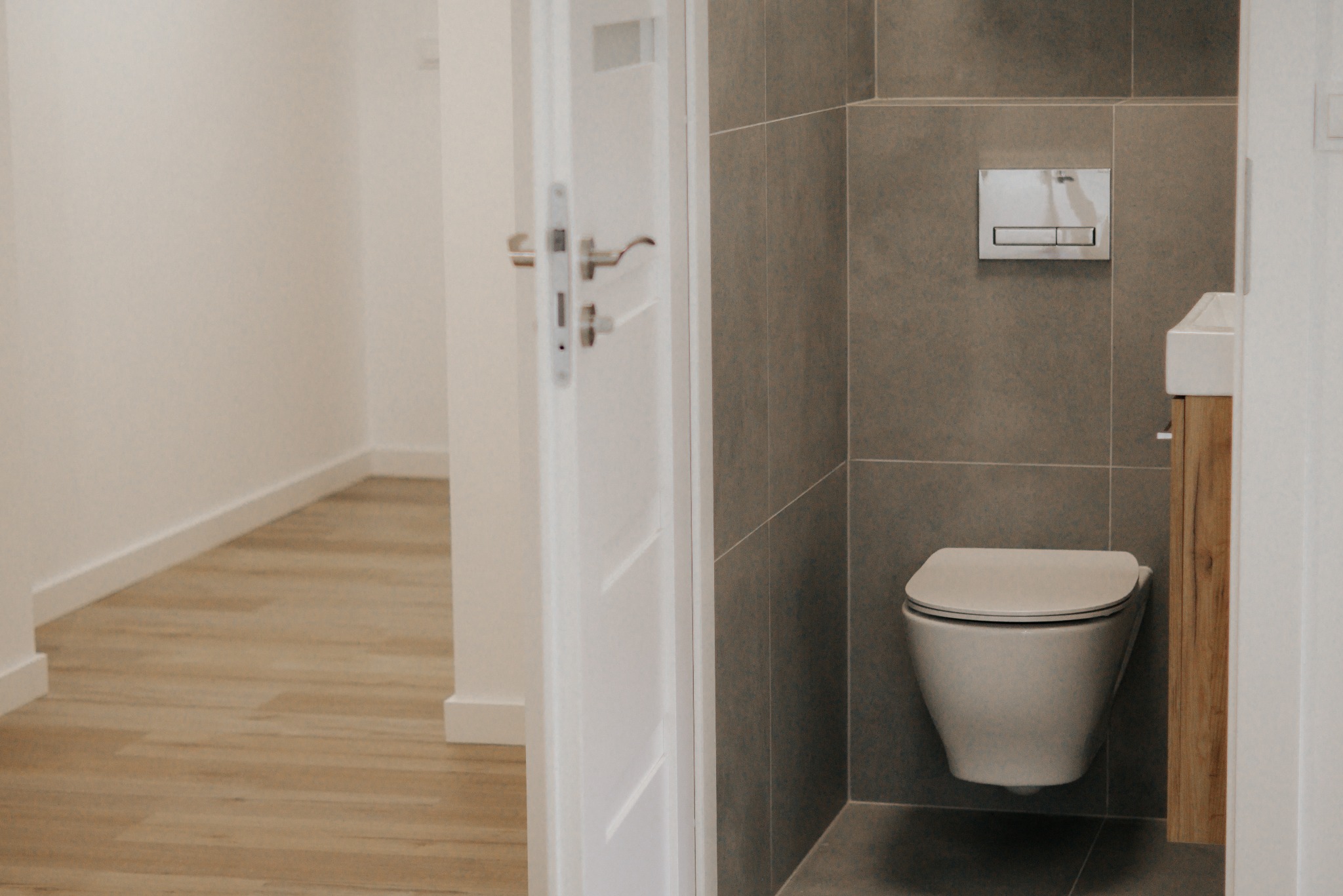 Nowoczesna łazienka z podwieszaną toaletą, szarą glazurą i drewnianą szafką pod umywalką. Widok zza otwartych, białych drzwi na parkiet w korytarzu.