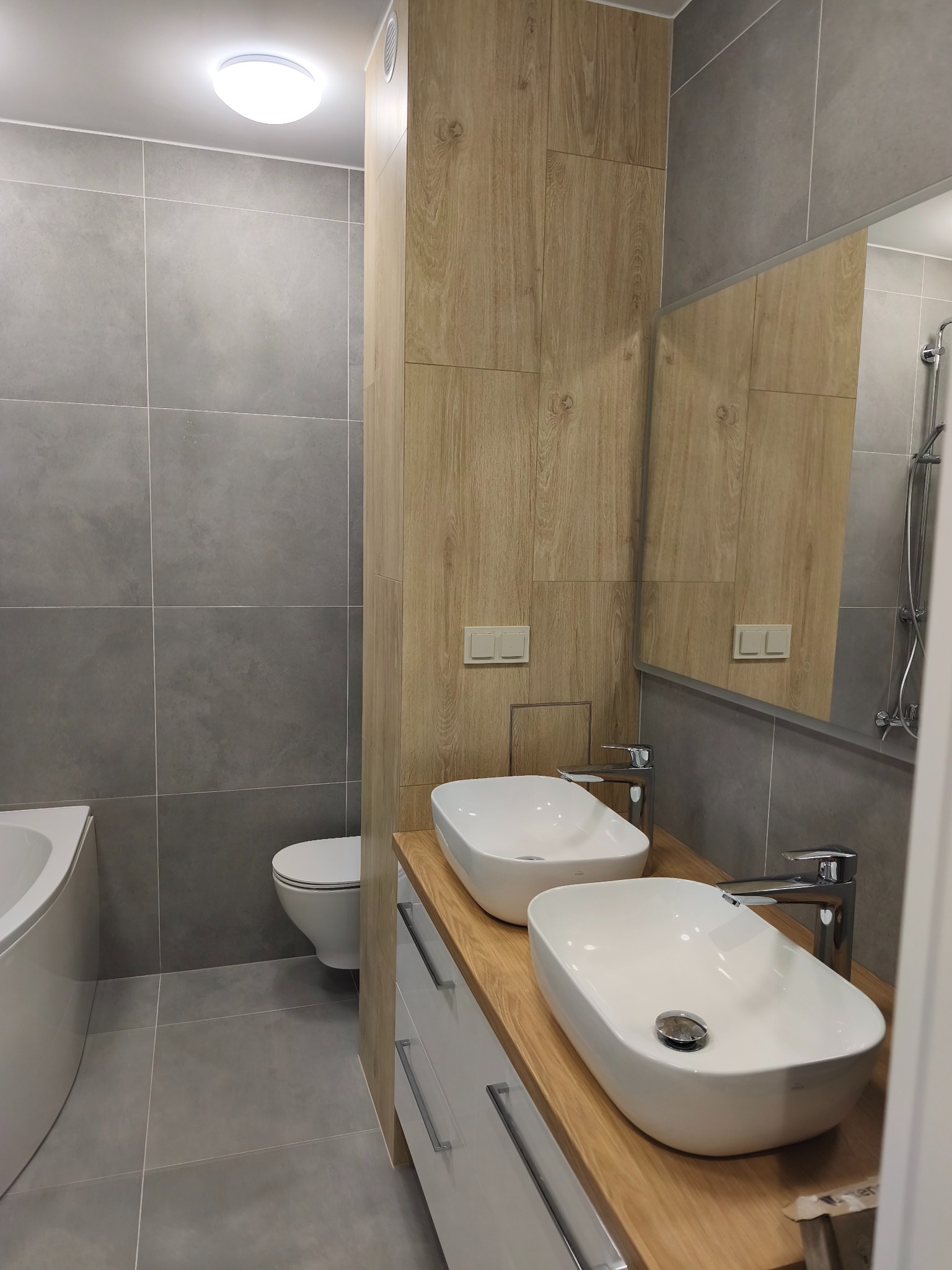 Nowoczesna łazienka po remoncie: szare płytki, drewnopodobne panele, dwie białe umywalki nablatowe i duże lustro. Minimalistyczny design z subtelnym oświetleniem.