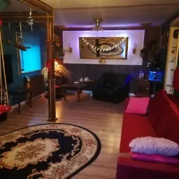 Salon w stylu eklektycznym z drewnianą podłogą, czerwonym aksamitnym narożnikiem i dywanem z kwiatowym wzorem. W tle widoczne lustro z dekoracyjnymi światełkami i akwarium z niebieskim oświetleniem.