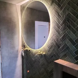 Ściana łazienki wyłożona ciemnozielonymi płytkami ułożonymi w jodełkę, okrągłe podświetlane lustro, złota armatura, fragment ściany w trakcie wykańczania.