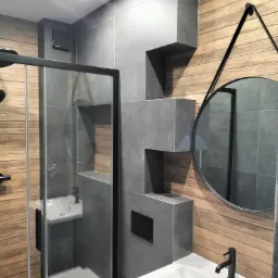 Nowoczesna łazienka z kabiną prysznicową typu walk-in, umywalką nablatową z czarną baterią i okrągłym lustrem na pasku, wykończona drewnopodobnymi płytkami i szarymi płytkami w geometrycznej formie.
