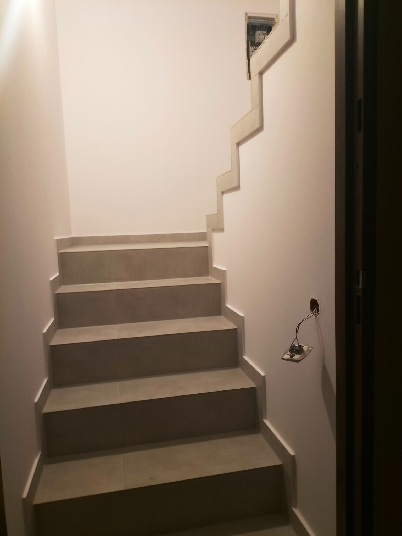 Schody wyłożone szarymi płytkami ceramicznymi, z widoczną instalacją elektryczną wystającą ze ściany i otwartą skrzynką elektryczną na górze ściany.