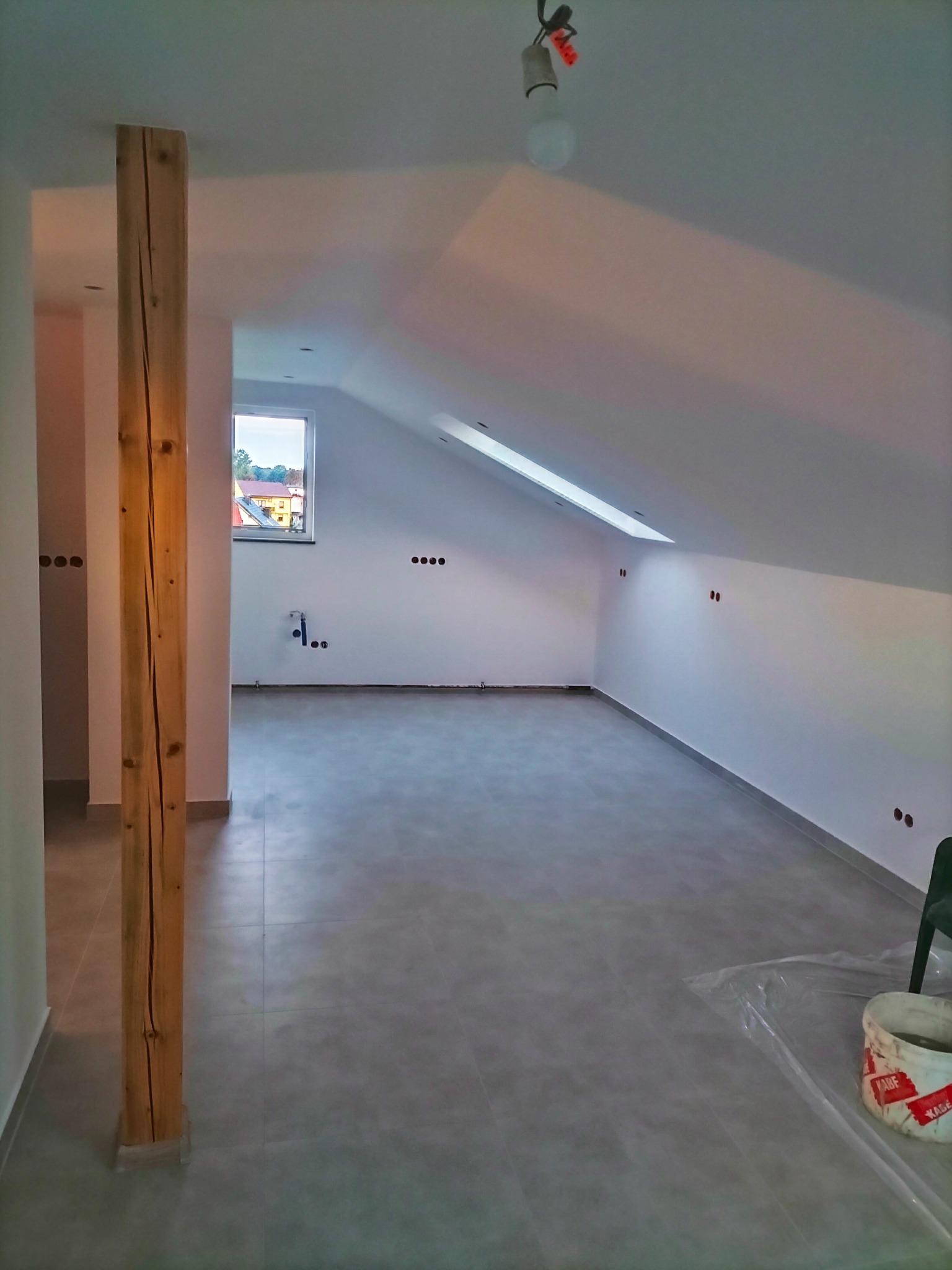 Poddasze po remoncie z szarą podłogą z płytek, białe ściany z widocznymi puszkami elektrycznymi, drewniany filar konstrukcyjny, okno dachowe i wisząca żarówka.