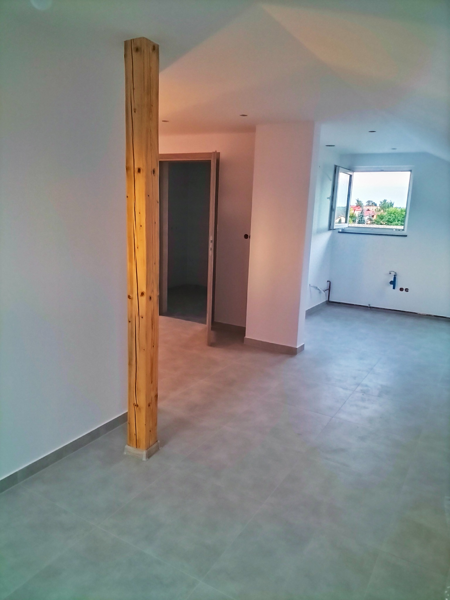 Jasne wnętrze po remoncie z szarą podłogą z płytek, drewnianym słupem konstrukcyjnym z widocznymi sękami i oknem z widokiem na zieleń.