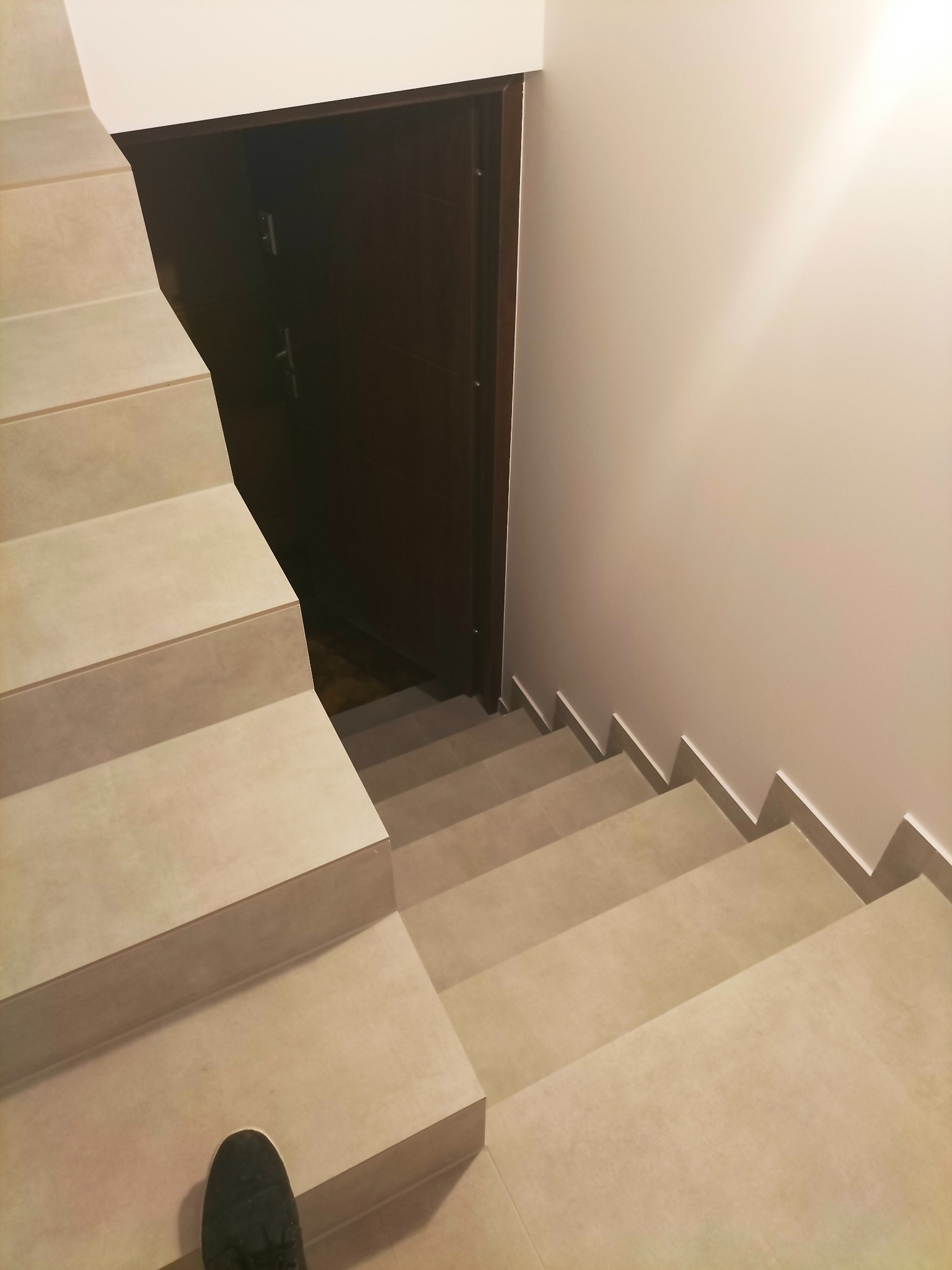 Wykończone schody z jasnych płytek ceramicznych prowadzące w dół, widoczny fragment buta na pierwszym planie i ciemne drzwi w tle.