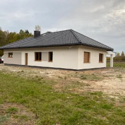 Nowoczesny dom jednorodzinny z ciemną dachówką i jasną elewacją, widok z zewnątrz na tle jesiennego krajobrazu i pochmurnego nieba, dwóch mężczyzn stoi przy samochodzie w tle.