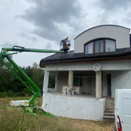 Pracownik na podnośniku koszowym maluje elewację domu jednorodzinnego, widoczne świeżo otynkowane ściany, częściowo zakryte przez zaparkowany biały samoch&oacute;d dostawczy.