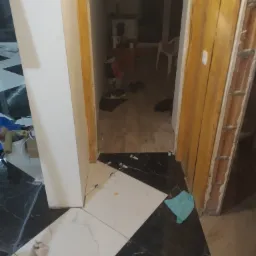 Biało-czarna podłoga z płytek ułożona w geometryczny wzór, widok na wnętrze przez otwarte drzwi z surową ościeżnicą, fragment niebieskiej szmatki na podłodze.