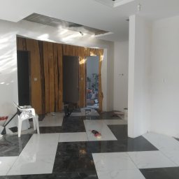 Maciej Jarosz - Wnętrze w trakcie prac: białe ściany, czarno-biała marmurowa podłoga w szachownicę, drewniana okładzina ścian z ukrytymi drzwiami, widoczne narzędzia i materiały budowlane, surowy sufit...