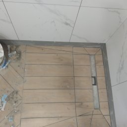 Maciej Jarosz - Niedokończona podłoga w łazience z drewnopodobnych płytek ułożonych wokół liniowego odpływu prysznicowego, widoczne krzyżyki dystansowe i wiadro z klejem.