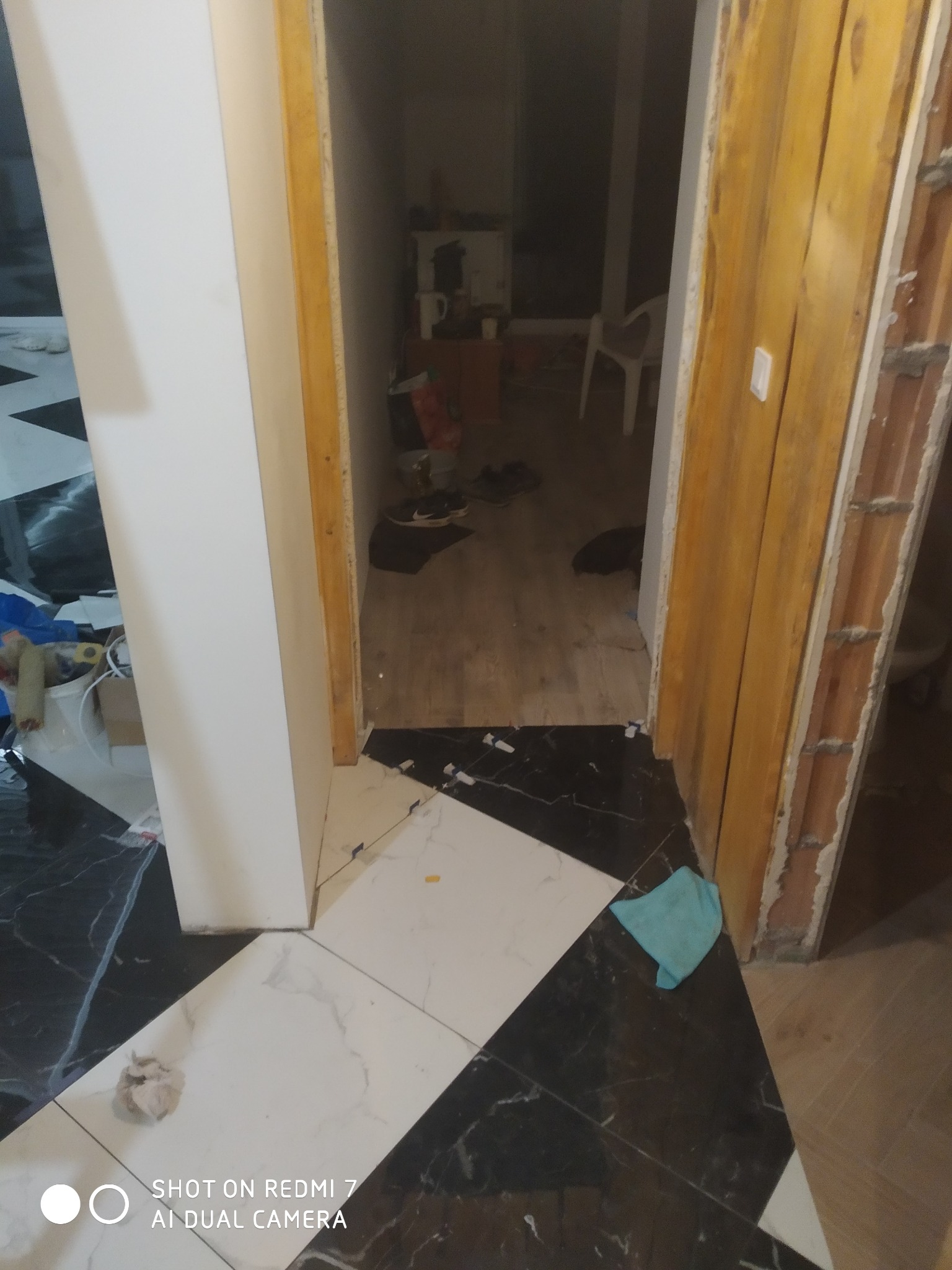 Biało-czarna podłoga z płytek ułożona w geometryczny wzór, widok na wnętrze przez otwarte drzwi z surową ościeżnicą, fragment niebieskiej szmatki na podłodze.