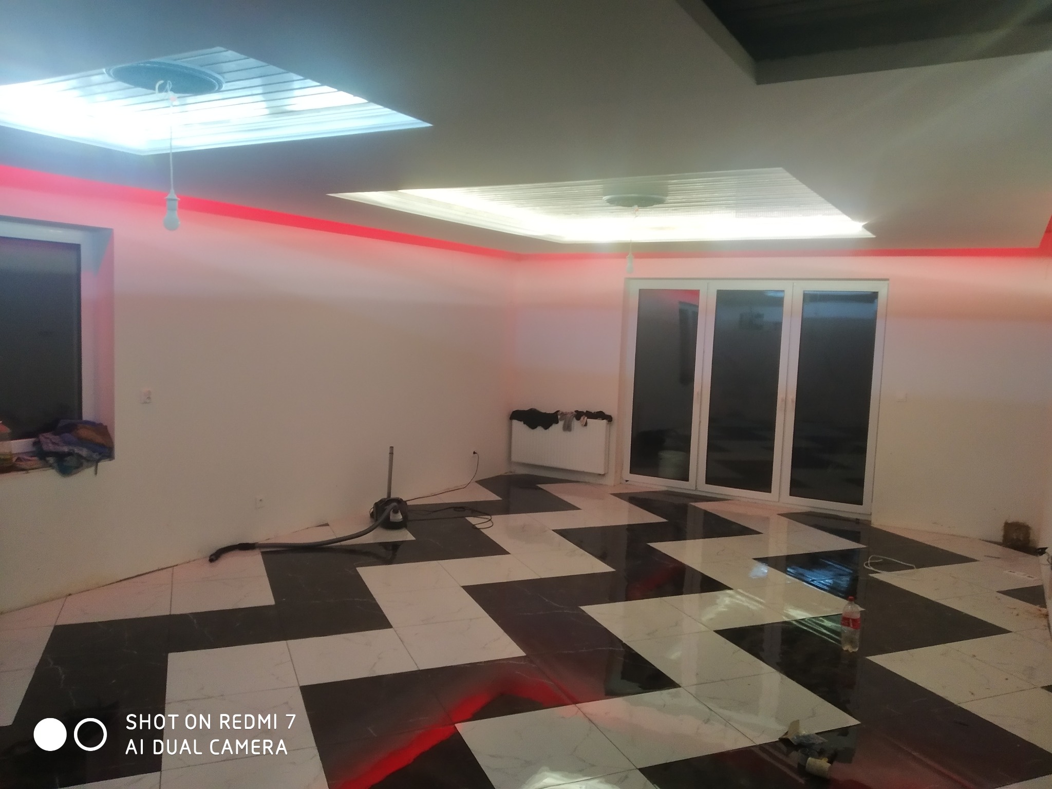 Pokój z ułożoną podłogą z czarno-białych płytek w zygzak, czerwone oświetlenie LED na suficie, okno i drzwi balkonowe, odkurzacz w rogu, widoczne ślady po remoncie.