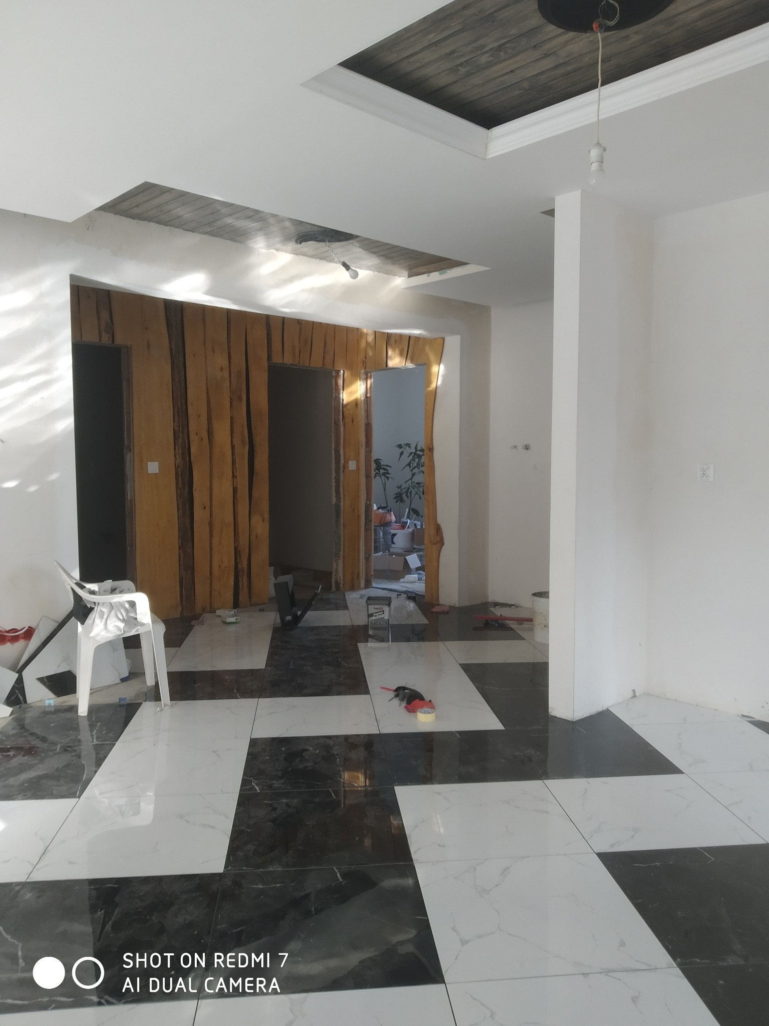 Wnętrze w trakcie prac: białe ściany, czarno-biała marmurowa podłoga w szachownicę, drewniana okładzina ścian z ukrytymi drzwiami, widoczne narzędzia i materiały budowlane, surowy sufit...