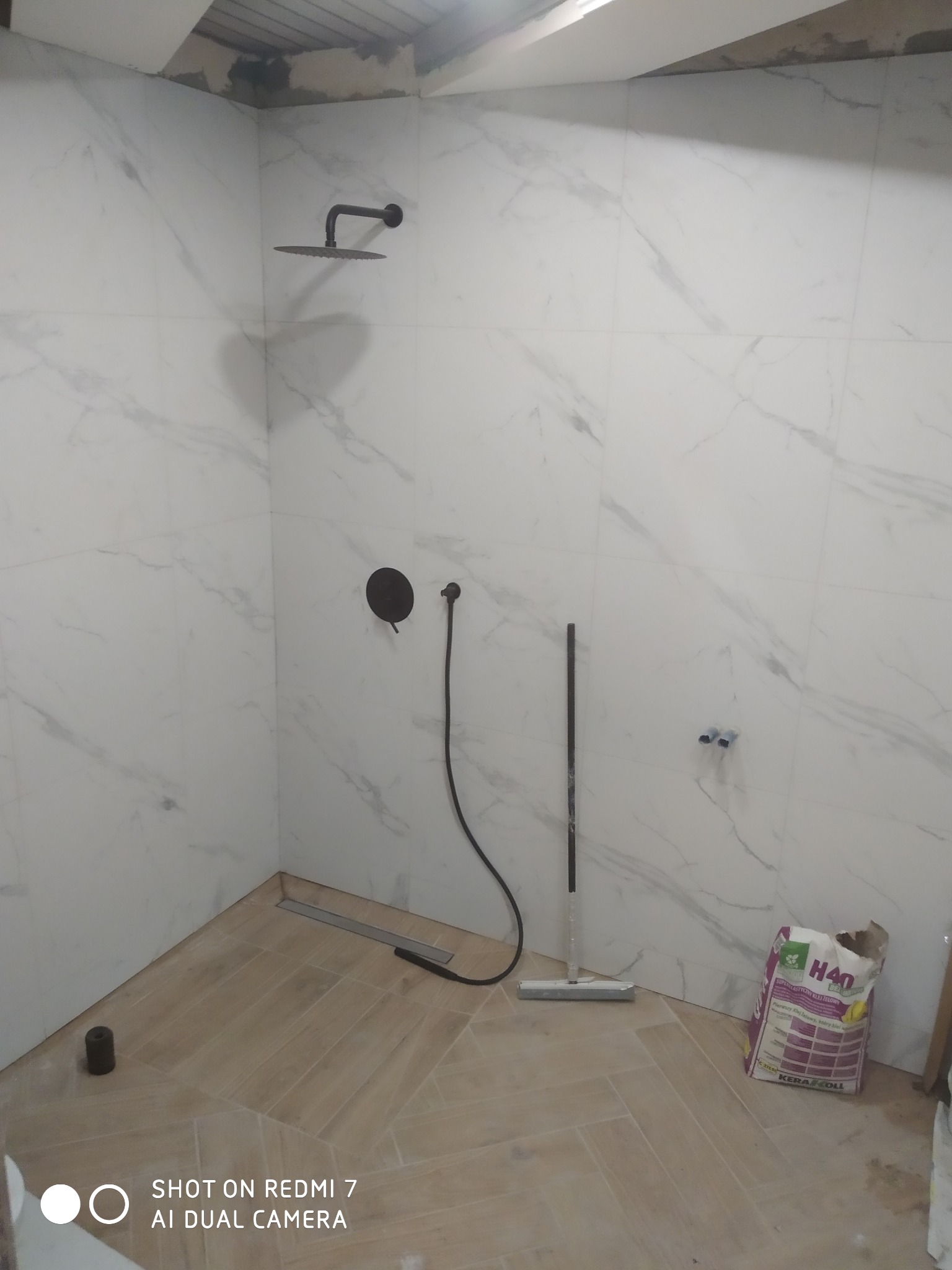 Łazienka w trakcie wykańczania: białe płytki imitujące marmur na ścianach, zamontowana czarna deszczownica i bateria, jasne płytki podłogowe ułożone w jodełkę, widoczny worek z klejem i narzędzia.
