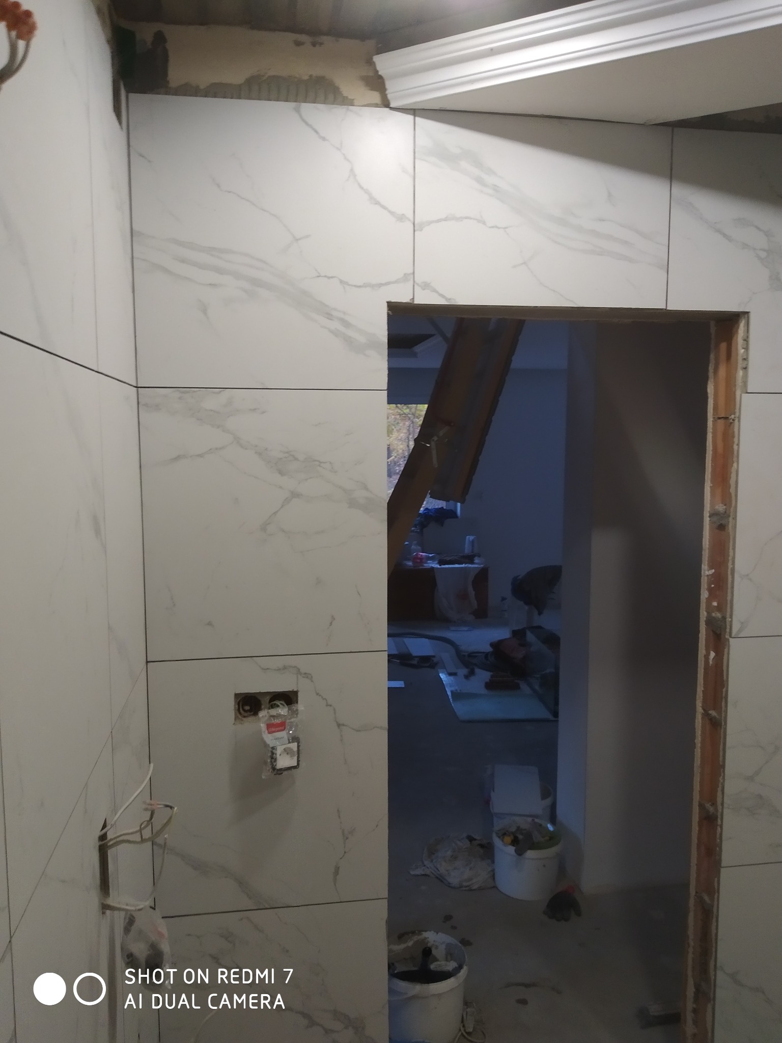 Łazienka w trakcie wykańczania: białe płytki imitujące marmur na ścianach, widoczne puszki elektryczne bez osprzętu, sufit z listwą dekoracyjną, bałagan w pomieszczeniu obok.