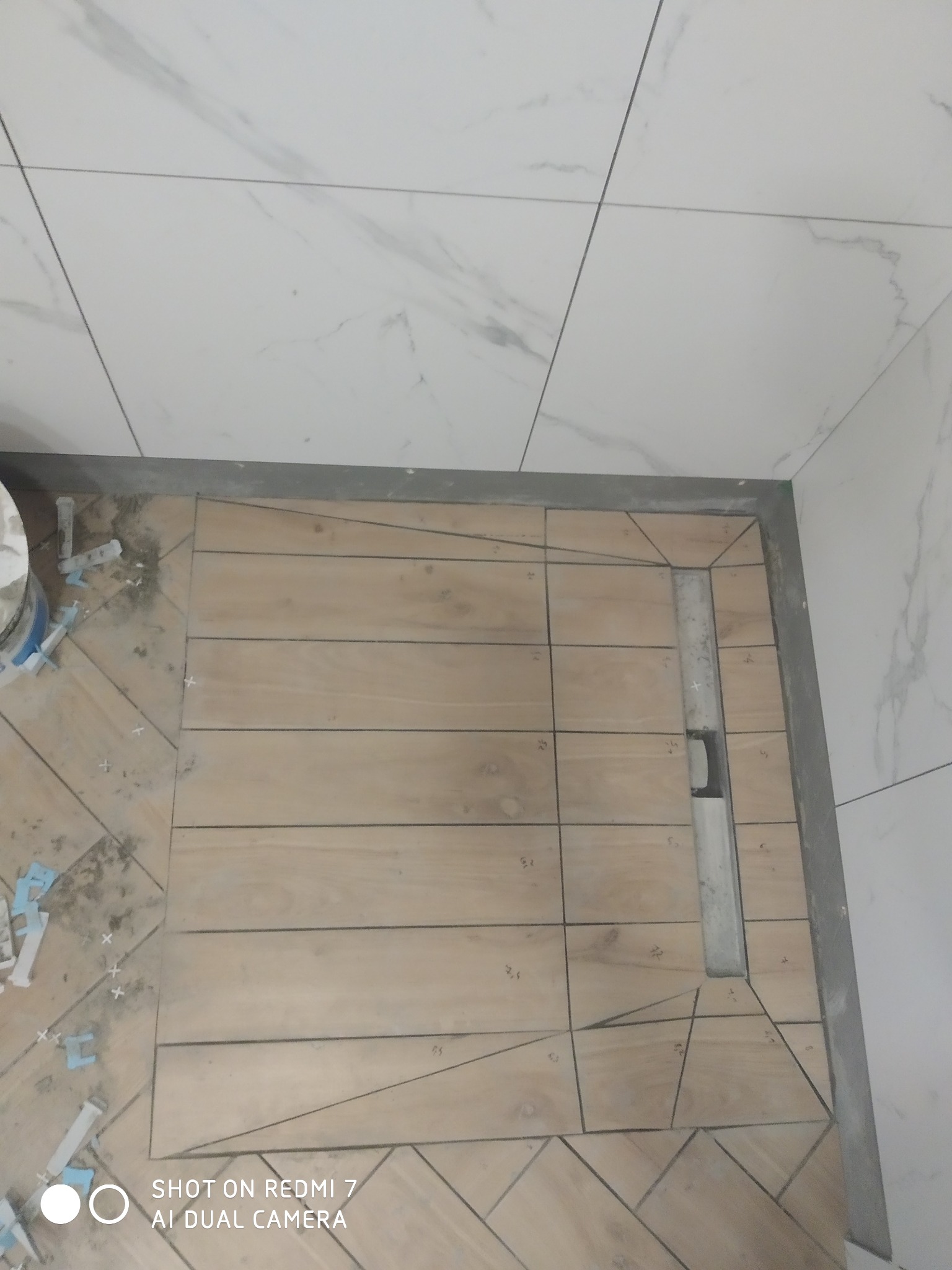 Niedokończona podłoga w łazience z drewnopodobnych płytek ułożonych wokół liniowego odpływu prysznicowego, widoczne krzyżyki dystansowe i wiadro z klejem.