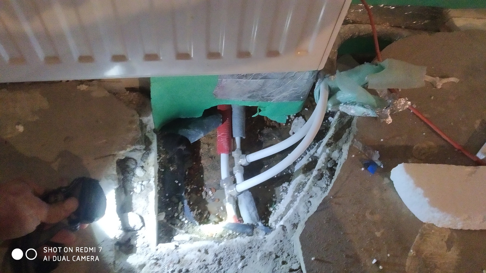 Niedokończona instalacja grzewcza przy grzejniku, widoczne rury z tworzywa sztucznego, izolacja z folii aluminiowej i zielonej pianki, oraz fragment podłogi w trakcie prac remontowych, oświetlona...