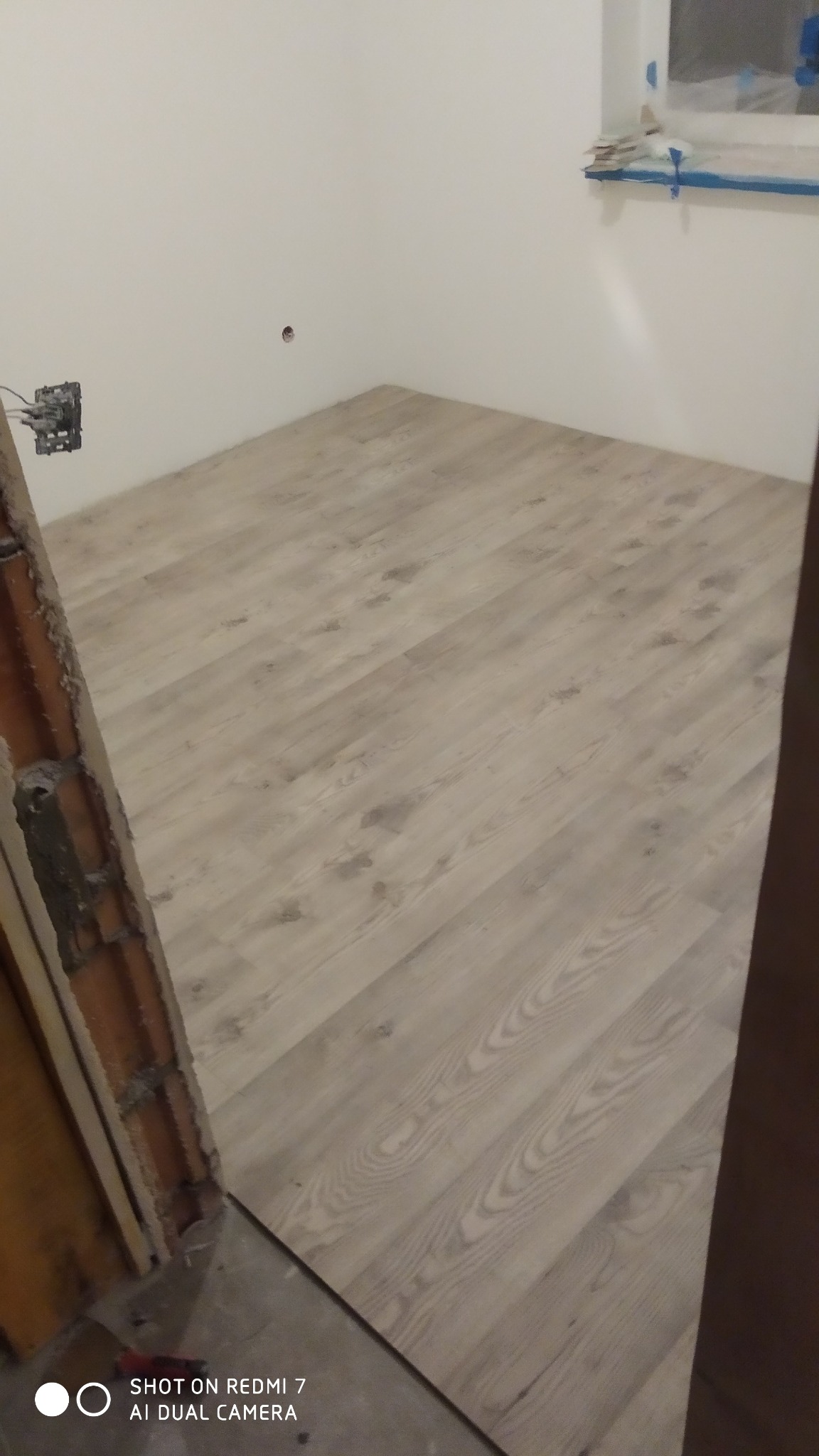 Nowo ułożone panele podłogowe w jasnym odcieniu drewna w pokoju z surowymi ścianami i odsłoniętą instalacją elektryczną, okno zabezpieczone folią malarską.