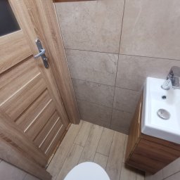 Rakso Efekt - Wykończona łazienka we Wrocławiu z drewnopodobnymi płytkami na podłodze i ścianach, widoczna umywalka z szafką i fragment drzwi.