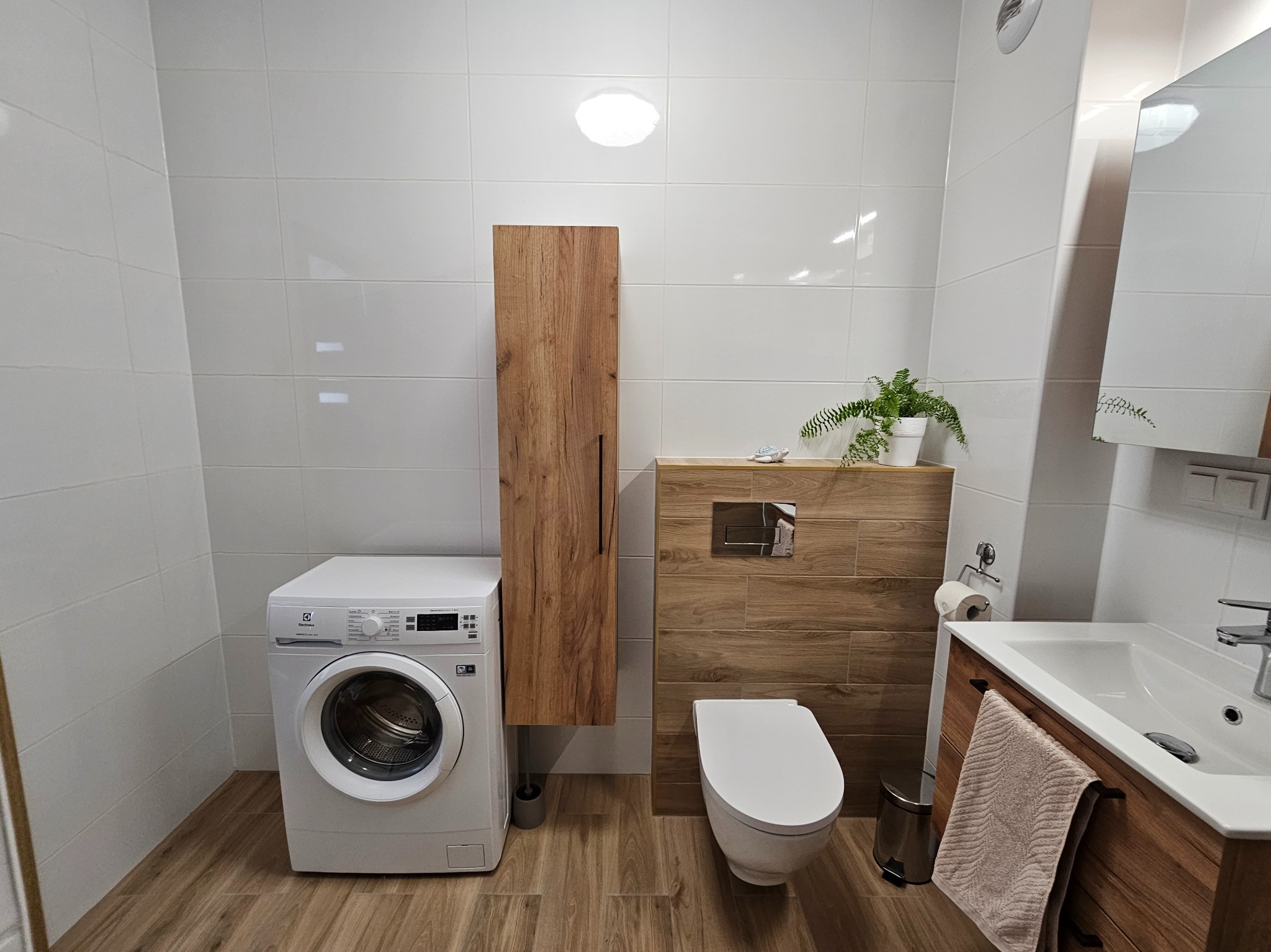 Nowoczesna łazienka z pralką Electrolux, toaletą, umywalką w szafce z drewnopodobnym wykończeniem i białymi, kwadratowymi płytkami na ścianach, oświetlona punktowym światłem.