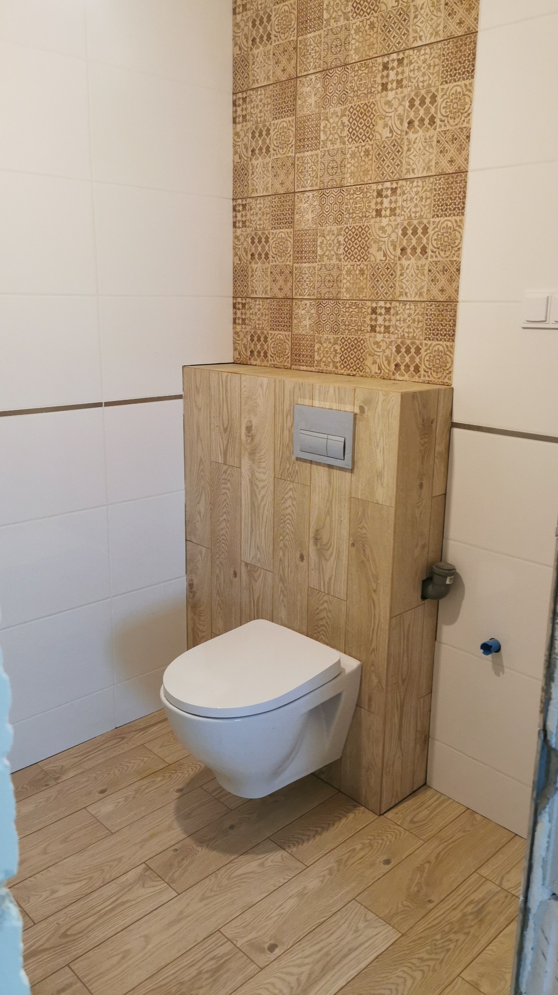 Nowoczesna toaleta podwieszana z zabudową z paneli imitujących drewno, wzorzyste płytki ceramiczne na ścianie w tle, jasne płytki na podłodze imitujące drewno, białe ściany.