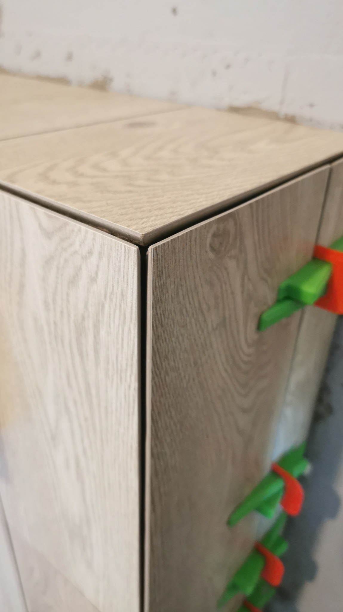 Fragment narożnika wykończonego płytkami imitującymi drewno, z widocznymi systemami poziomowania płytek (klipsy i kliny) w kolorach zielonym i pomarańczowym.