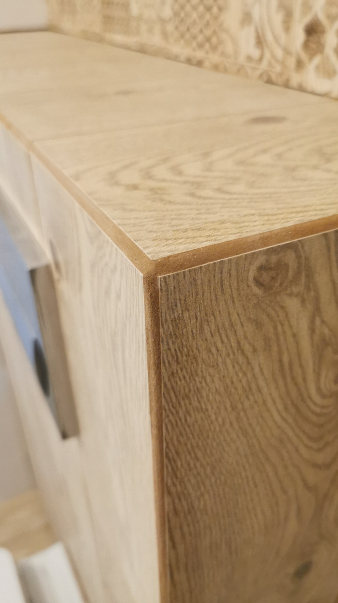 Szczegółowe ujęcie narożnika szafki łazienkowej z płyty imitującej jasne drewno, z widoczną krawędzią wykończeniową w kolorze naturalnego drewna i fragmentem dekoracyjnej płytki na ścianie w tle.