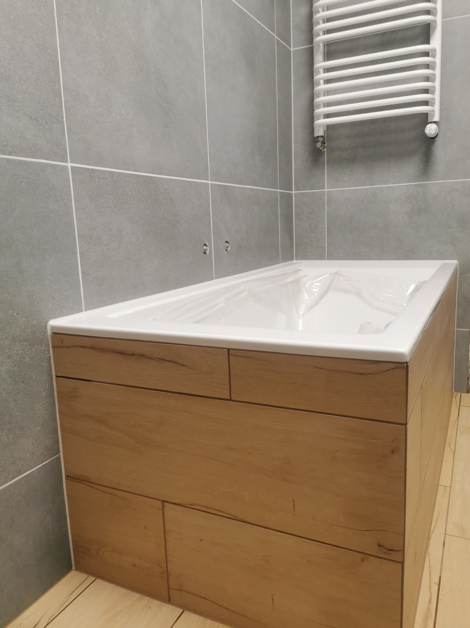 Nowa wanna obudowana panelami imitującymi drewno, zabezpieczona folią ochronną, na tle szarych płytek i białego grzejnika drabinkowego w łazience.