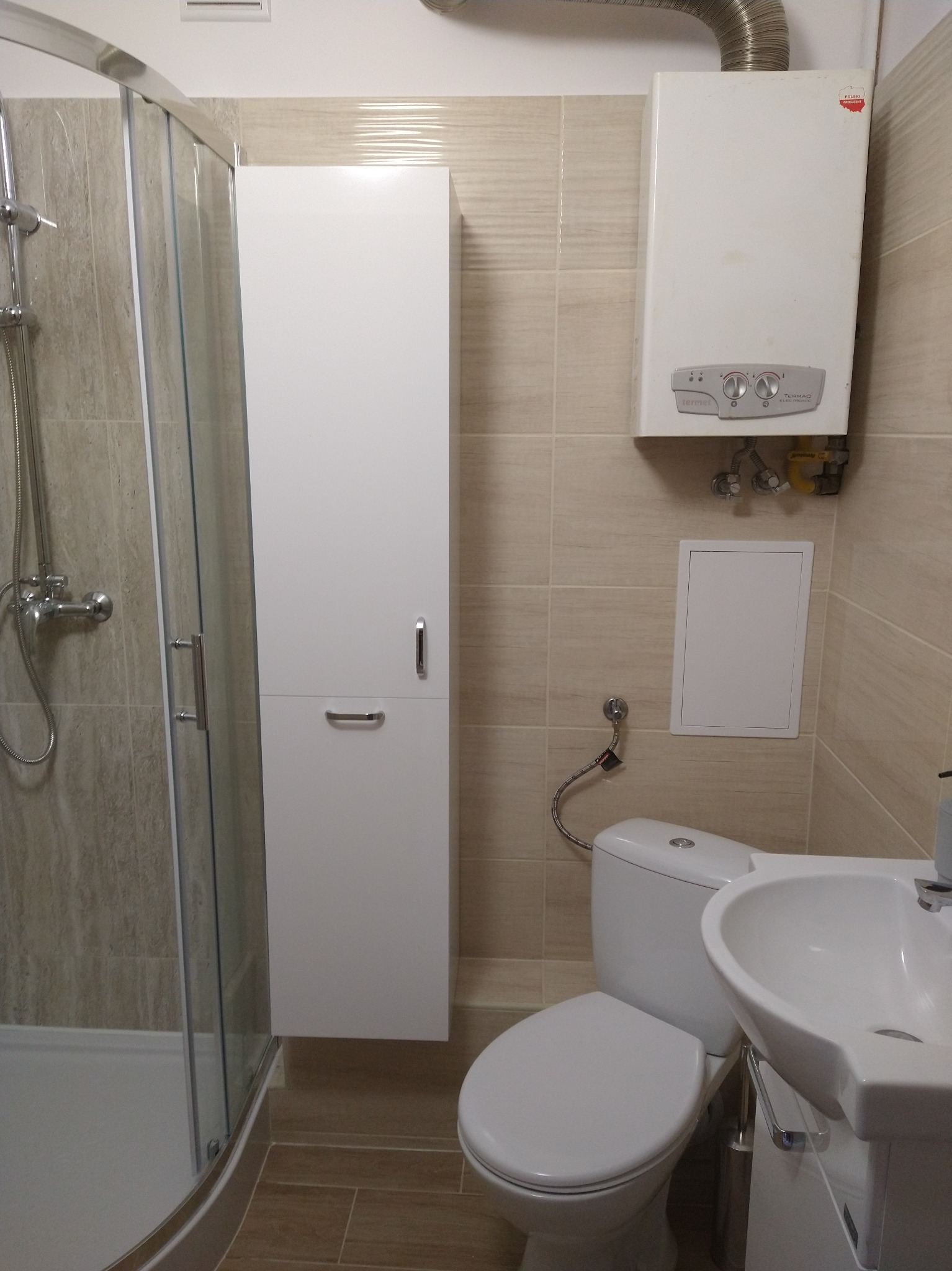 Wykończona łazienka z kabiną prysznicową z chromowanymi detalami, białą szafką wiszącą, toaletą i umywalką. Na ścianie beżowe płytki oraz piec gazowy.