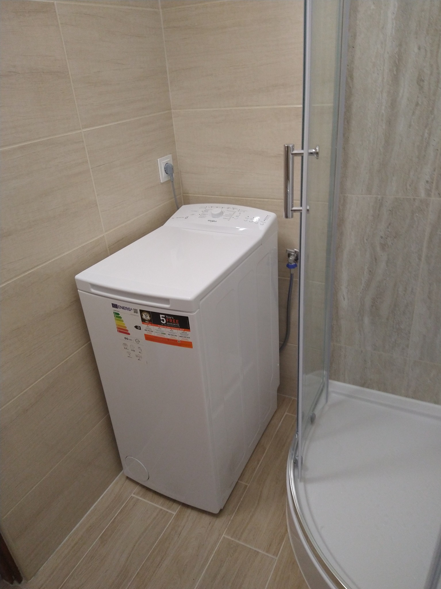 Biała pralka w narożniku łazienki z beżowymi płytkami, obok kabina prysznicowa z przezroczystymi drzwiami.