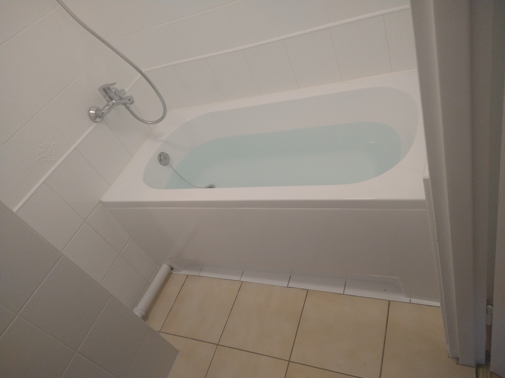 Biała wanna wypełniona wodą, widok z góry, z chromowaną baterią i słuchawką prysznicową na białej, kafelkowanej ścianie, z beżową podłogą wyłożoną kwadratowymi płytkami.