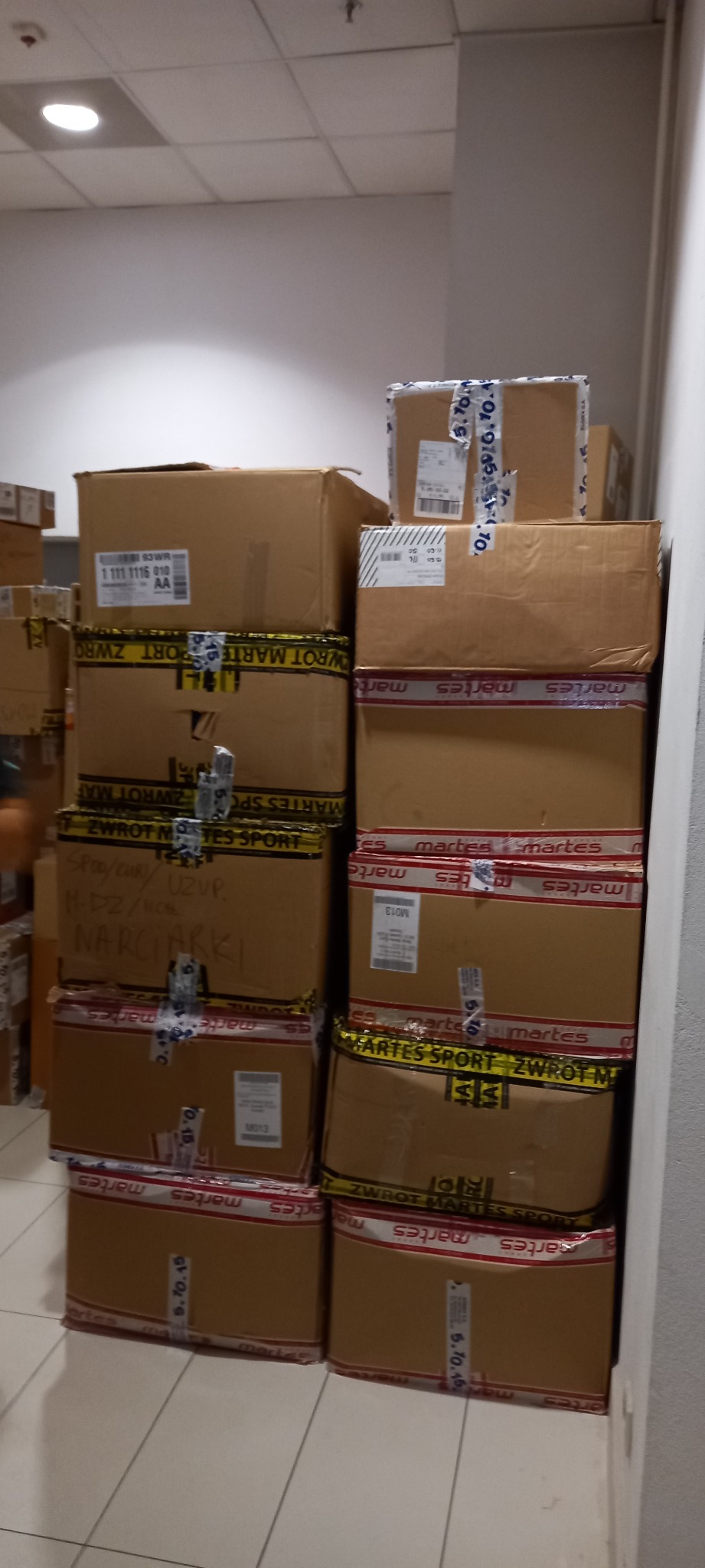 Stos kartonowych pudeł oklejonych taśmą, niektóre z napisem 'ZWROT MARTES SPORT' i adresem, ustawionych przy białej ścianie w magazynie lub biurze.