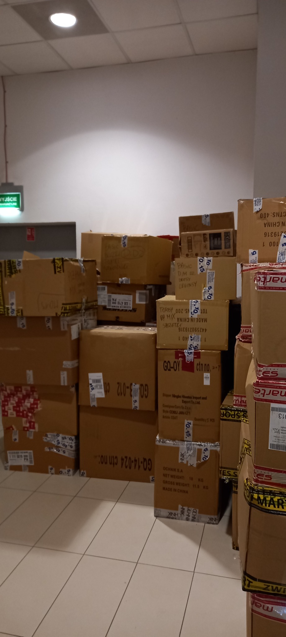 Stosy kartonowych pudeł z etykietami i taśmą, ułożone w magazynie lub hali, widoczny znak wyjścia ewakuacyjnego.