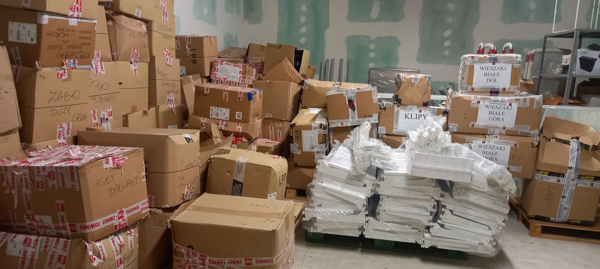 Stos kartonowych pudeł z napisem 'Zwrot Towaru' i oznaczeniami zawartości, takich jak 'Wieszaki', 'Klippy', 'Żabki', 'Igły', składowanych w magazynie z surowymi ścianami.