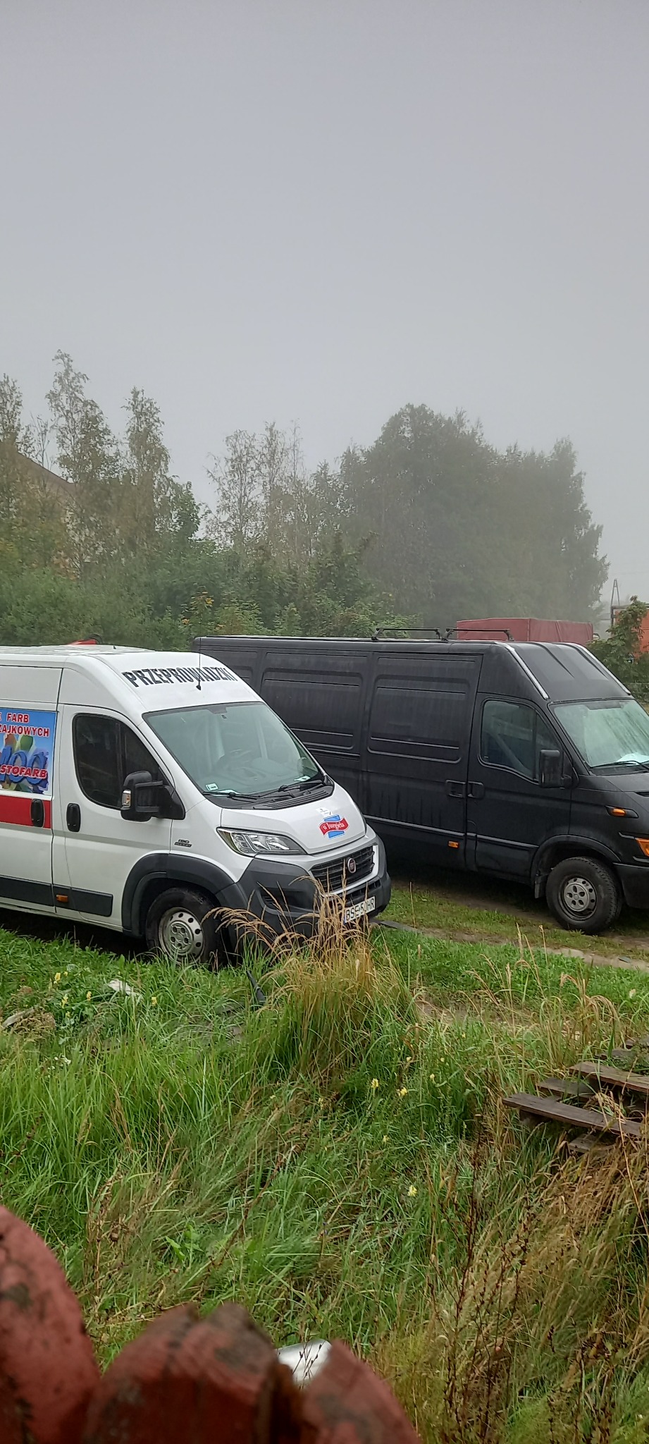 Dwa busy: biały z napisem na dachu i kolorową grafiką na boku oraz czarny, stoją na trawiastym terenie w pochmurny dzień.