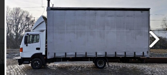 Biała ciężarówka z plandeką zaparkowana na brukowanej powierzchni, widoczna z boku na tle torów kolejowych i drzew bez liści.